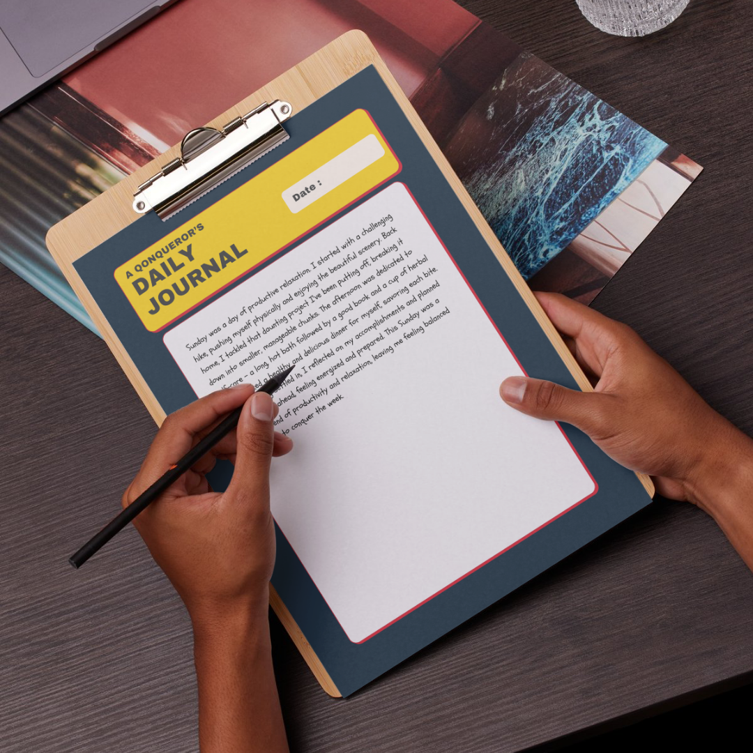 A person with brown skin and dark hair sits at a wooden desk, holding a black pen and writing in a journal on a clipboard. The clipboard has a yellow header that says "A Conqueror's Daily Journal" and a space for the date. There is a laptop and other papers on the desk.